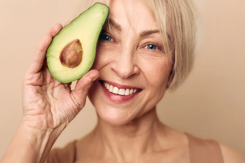 Smiling mature woman with clear skin holds fresh avocado half near her face on beige background. Concept for healthy lifestyle, natural beauty, skincare, nutrition, and wellness.