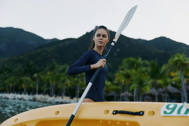Athletic kayaker stands aboard a bright yellow kayak, gripping a paddle as she prepares to glide across calm waters amid tropical scenery and distant forested mountains.