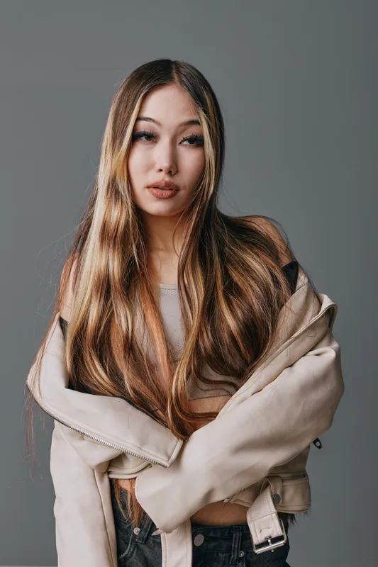 Young woman with long hair posing in neutral jacket against gray background for fashion portrait, beauty, style and confident expression in studio lighting.