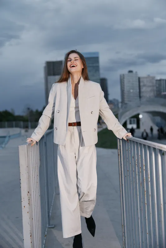 Confident woman in beige business suit laughing outdoors in urban setting with modern buildings and cloudy sky background