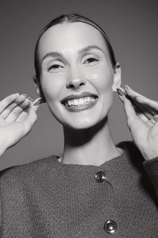 Happy Caucasian woman with fair skin smiling and holding earrings near her ears. Studio portrait with soft lighting and minimalistic style, concept of beauty and fashion.