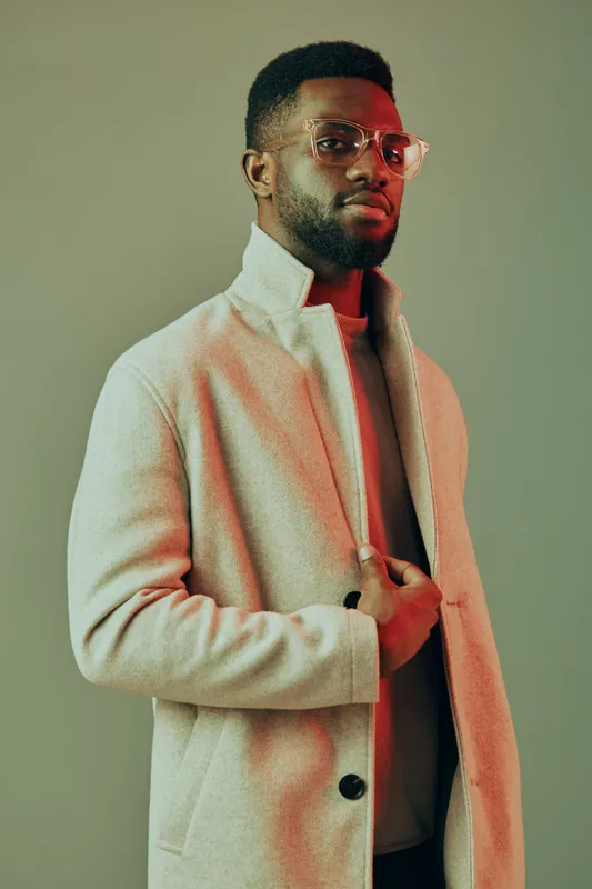 Stylish young man in a light coat and glasses posing confidently against a muted background, showcasing modern fashion and elegance