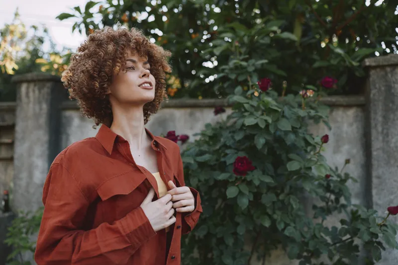 Cheerful woman outdoors, wearing a rust shirt, head tilted, eyes closed in a gentle smile, soft sunlight, natural colors, leafy garden backdrop, relaxed lifestyle moment