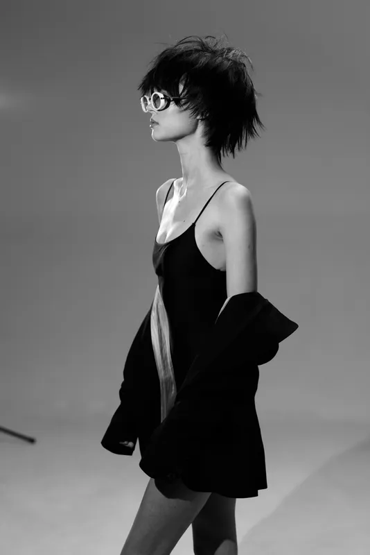 Black and white portrait of a stylish woman wearing sunglasses and a black dress with a jacket draped over her arms.