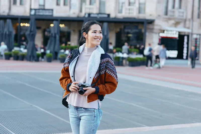 Woman with camera in hand smiles while strolling on a city square, wearing a cozy patterned jacket and jeans, capturing candid moments of urban life and everyday photography in daylight