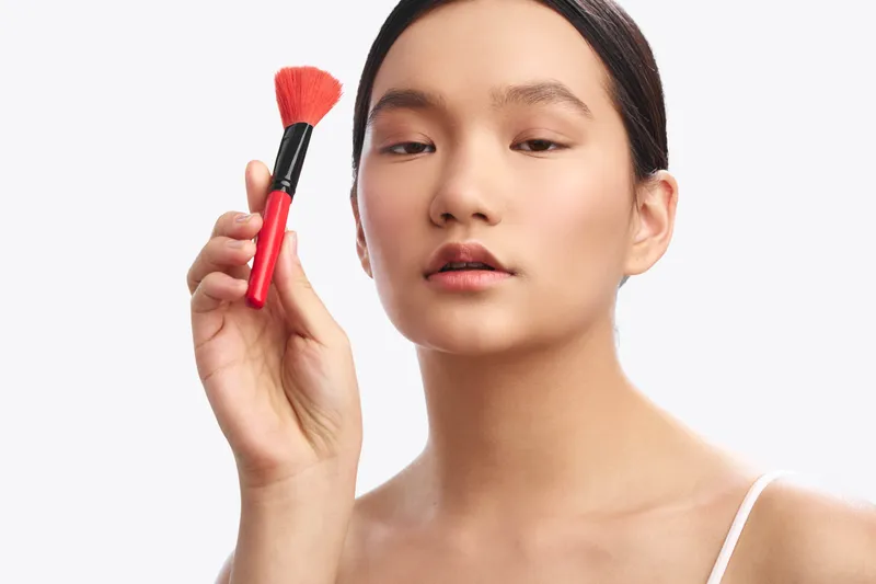 asian woman applies makeup with a brush in a bright studio, showing a calm, focused expression. the clean composition highlights beauty routine and contemporary style, creating a polished, confident