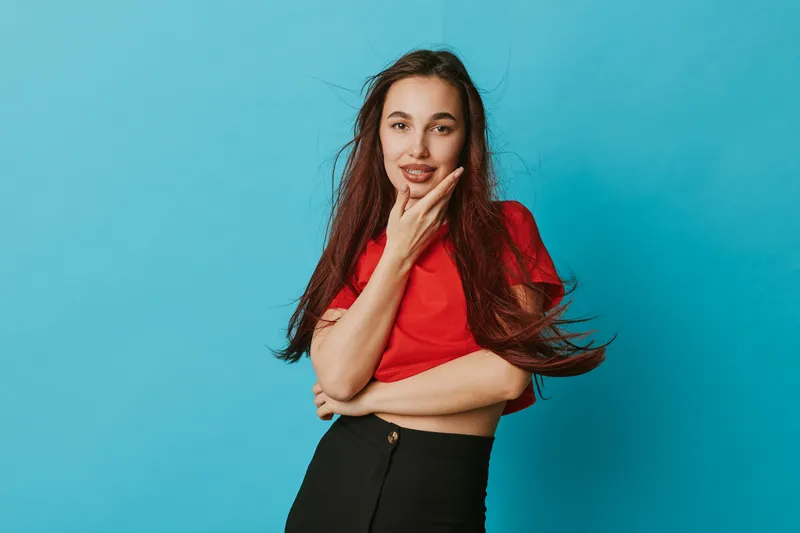 Young woman with long hair wearing red shirt and black pants posing with hand on chin against blue background. Female model looking confident and thoughtful in studio portrait.