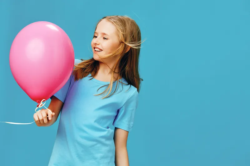 Young girl with long blond hair joyfully holding a pink balloon against a vibrant blue background, capturing happiness and childhood innocence in a playful moment