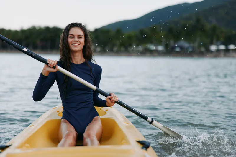 Young woman paddles a yellow kayak on calm water, wearing a dark blue wetsuit, smiling as she rows, with splashes and a distant shoreline under an open sky, surrounded by gentle breeze and soft light.