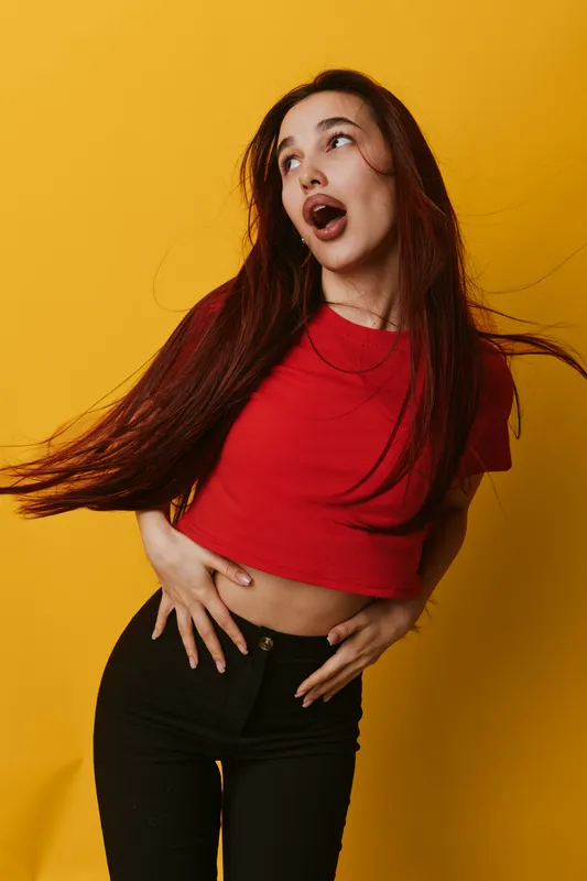 Woman with long hair wearing red crop top and black pants posing expressively on yellow background. Female model with open mouth and dynamic hair movement for fashion or lifestyle.
