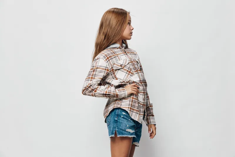Girl standing in profile pose wearing plaid shirt and denim shorts against white background. Casual outfit and confident posture with hand on hip for fashion look.