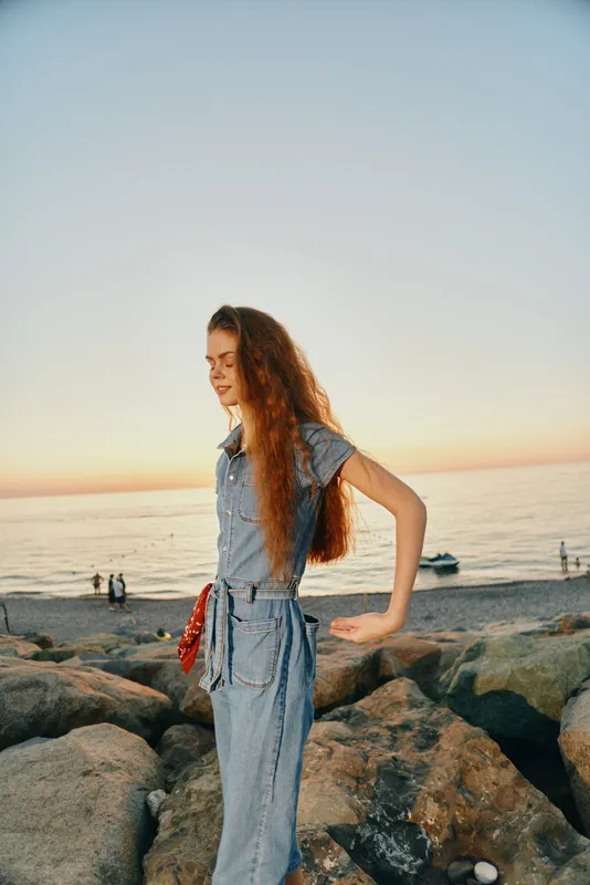 lifestyle woman in modern boho-western denim street style standing on rocks by the sea with warm film color sunset lighting
