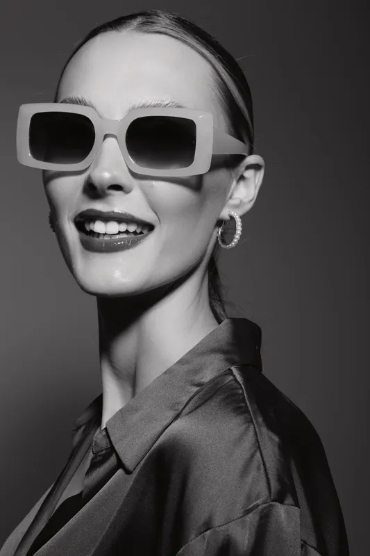 Smiling white woman with pale skin wearing large sunglasses and hoop earrings in a black satin shirt. Studio portrait with high contrast lighting for fashion and style concept.