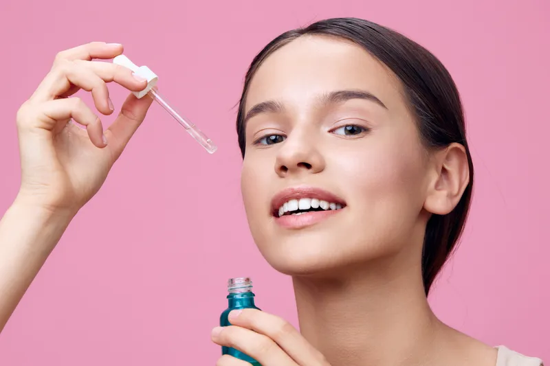 Young woman applying skincare serum, smiling, on a pink background, promoting healthy skin and beauty concepts through vibrant colors and freshness