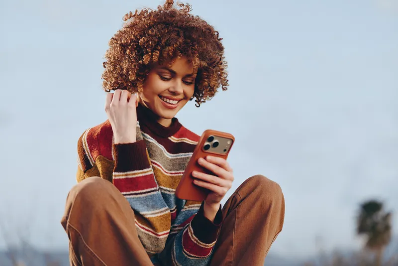 Smiling woman with curly hair using smartphone outdoors, casual lifestyle portrait against soft sky background, seated relaxed mood, self confidence, friendly communication today