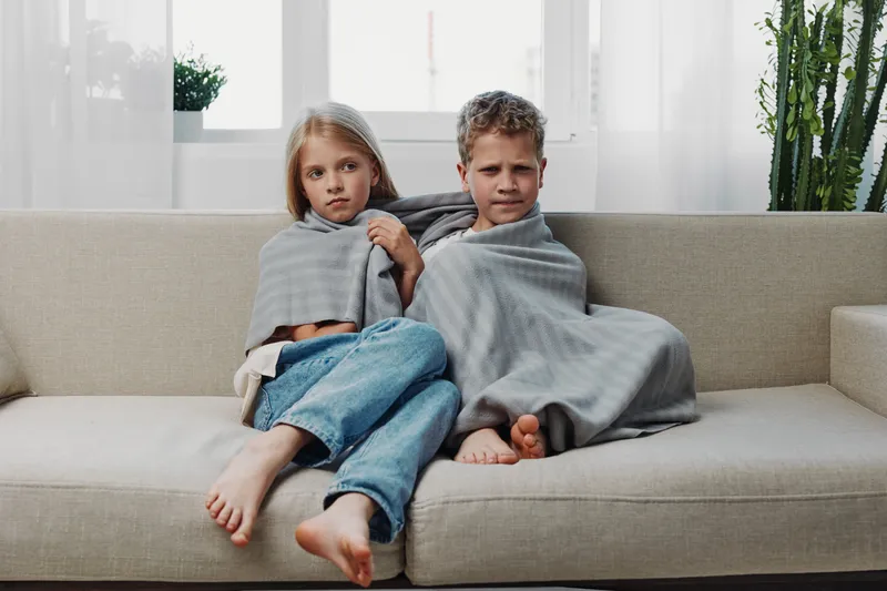Two children wrapped in gray blankets sit barefoot on a beige sofa in a bright living room. The kids look serious, conveying comfort and quiet moments at home during cold weather.