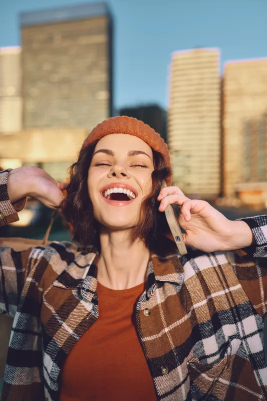 Woman smile phone urban beanie golden hour candid woman smiling on phone against city skyline at golden hour, plaid jacket and beanie, authenticity, candid lifestyle, mindful living and emotional