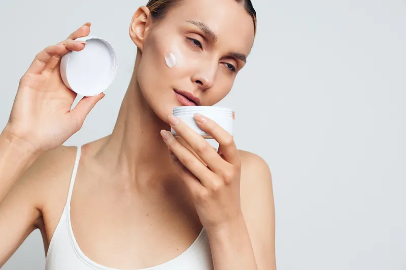 Young woman applying white face cream on cheek with closed eyes, holding open jar in hand against white background, concept of skincare, beauty routine, and self-care.