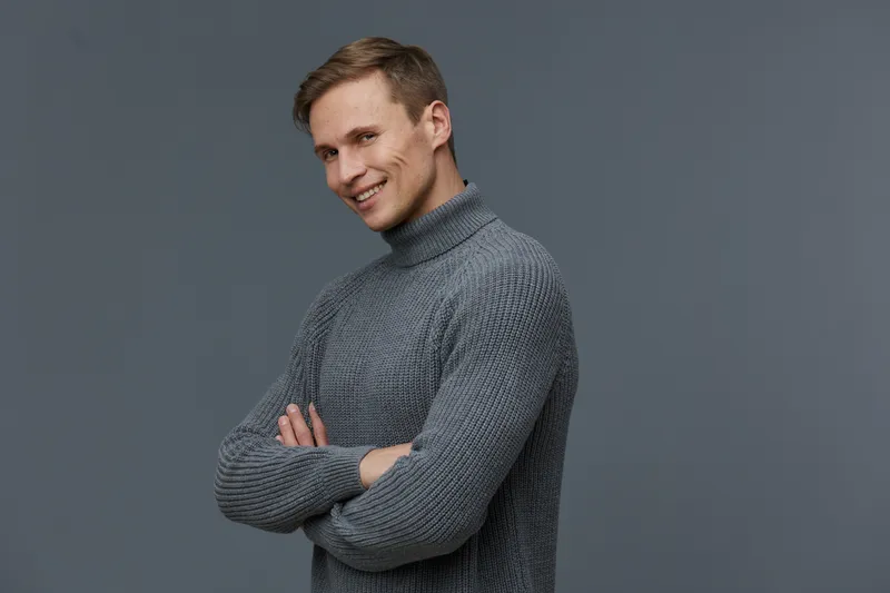Smiling young man with short hair wearing cozy gray turtleneck sweater looking confidently at camera on gray studio background. Casual fashion lifestyle concept.