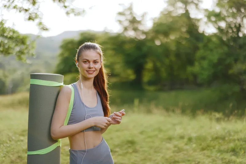 Young fit woman in sportswear with yoga mat, smiling confidently outdoors in a natural setting, embodying health and fitness