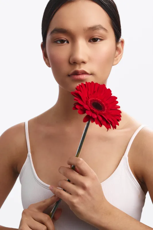 Asian woman holds a bright red flower close to her face in a minimalist studio portrait. She wears a white tank top, calm expression, and soft studio lighting.
