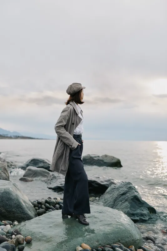 A woman in a long coat and hat stands on rocks by the sea, looking towards the horizon under a cloudy sky. Concept of solitude, nature, and contemplation at the coast.