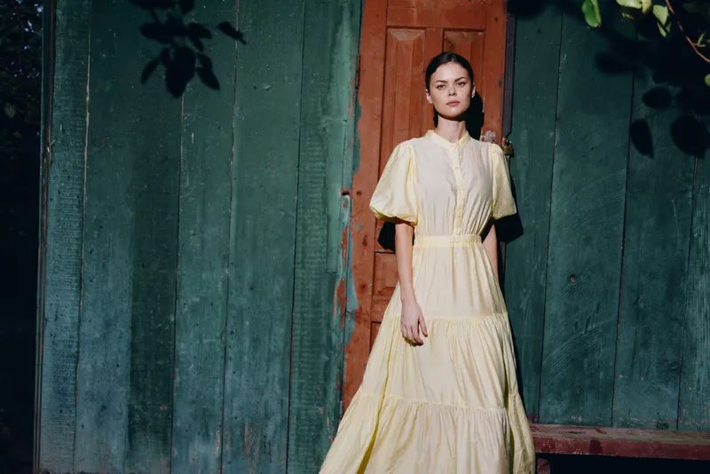 Woman portrait standing in vintage yellow dress by rustic doorway with sunlight authenticity inclusivity and natural expression fashion photography calm mood country soft styling