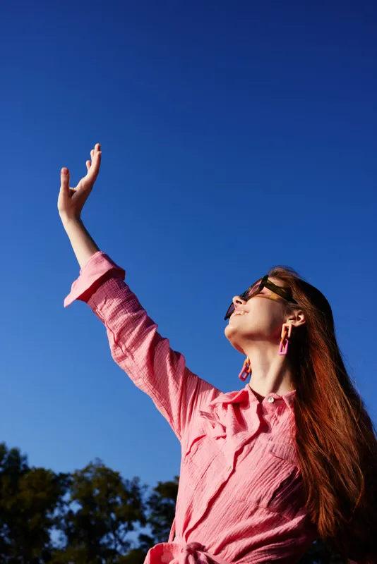 A young woman with long hair reaching out towards the blue sky, wearing a bright pink shirt and stylish sunglasses, expressing joy and freedom in a vibrant summer setting