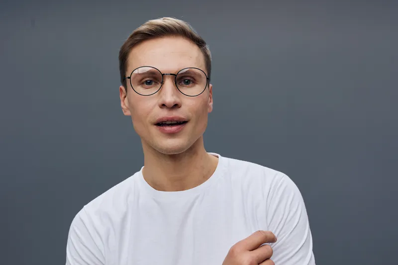 Close-up of a confident young Caucasian man with glasses wearing a white t-shirt, looking directly at camera against a plain gray background. Studio portrait with a professional and approachable vibe.