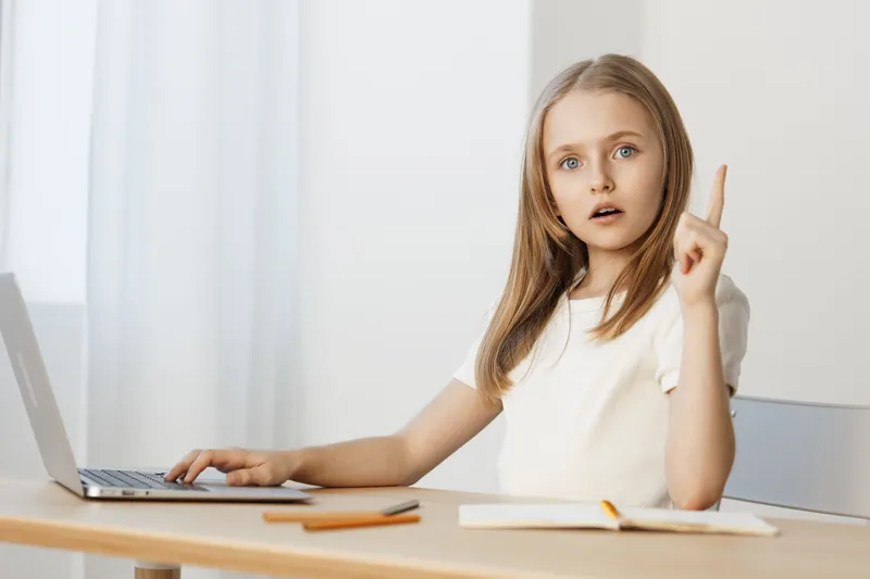 Thoughtful girl with long hair sitting at a desk, raising her finger in a moment of inspiration, using a laptop and notebook, focused on her work and ideas