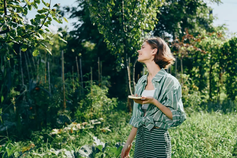 A woman stands in a sunlit garden, holding a plate as she admires fresh greenery and outdoor life.
