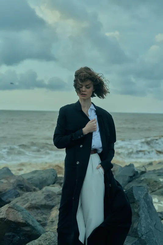 Fashion, woman, coat, ocean, wind, coastline, outdoor, styling showcased as a model stands by the sea in a long dark coat over light trousers, capturing a moody, cinematic moment under a cloudy sky
