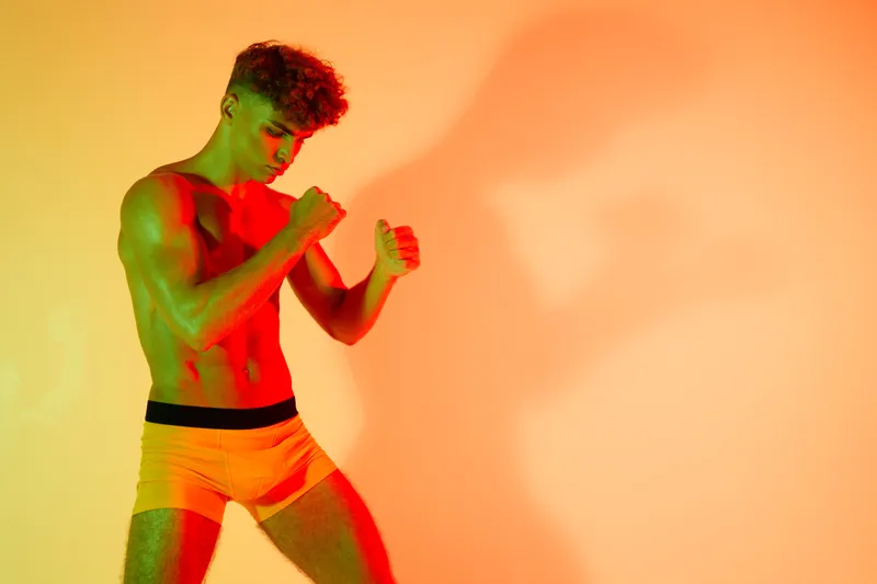 Young man with muscular body posing in boxer briefs with colorful lighting and shadow on wall for fitness, health and athletic lifestyle concept.