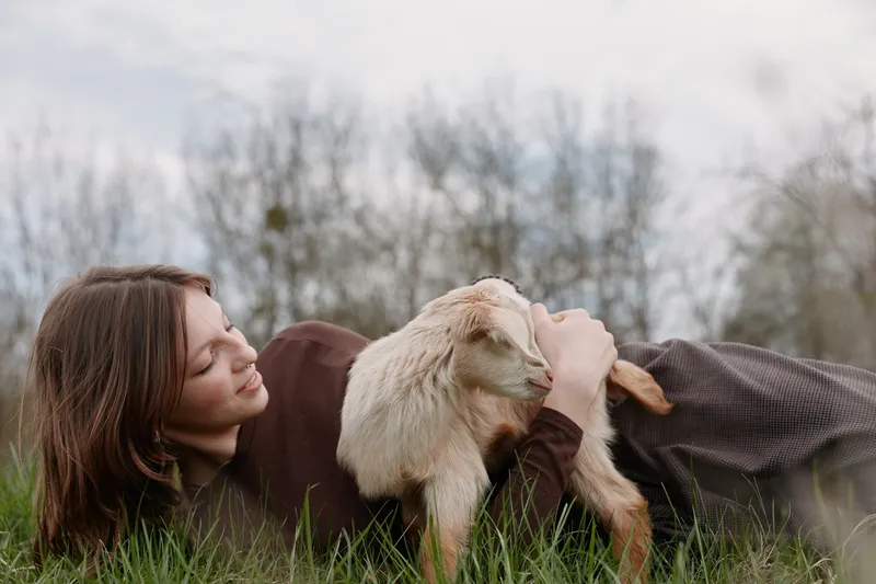 Relaxed woman lying in grass beside a curious goat kid, sharing a gentle moment in a rural setting, showcasing bond with animals and nature, peaceful outdoor scene with soft light.