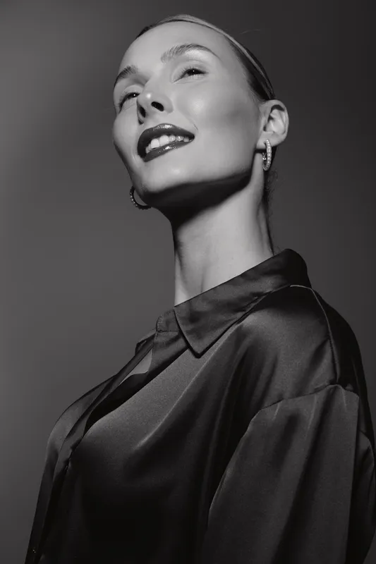 Close-up portrait of a smiling caucasian woman with fair skin wearing a dark satin blouse and earrings. Studio lighting creates elegant and confident mood in black and white photo.