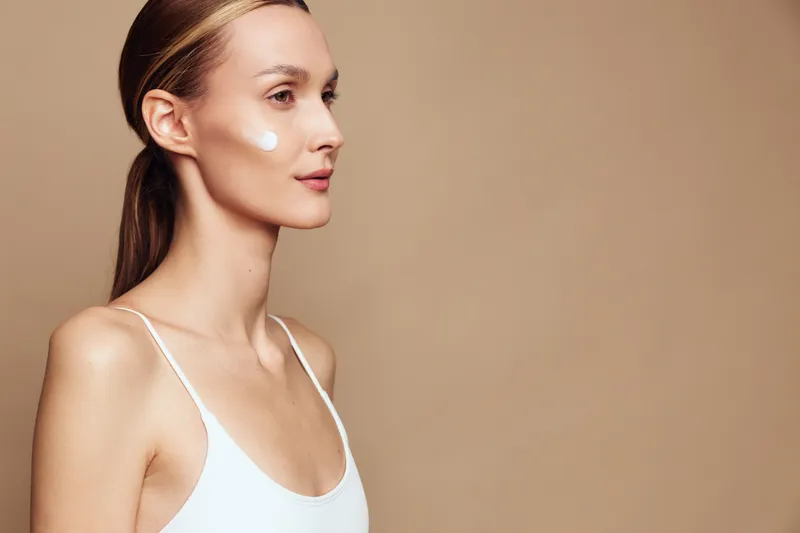 Young woman with clear skin applying cream on face, side profile, natural makeup, soft lighting, neutral background, skincare routine, focused expression