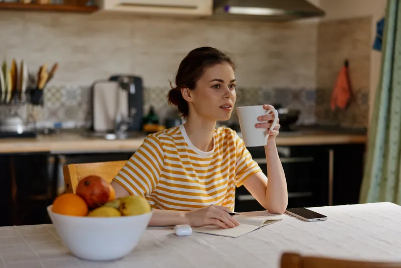 Casual woman sits at a kitchen table sipping coffee while writing notes, home lifestyle scene with fruit bowl nearby, calm focused mood, everyday moment at home