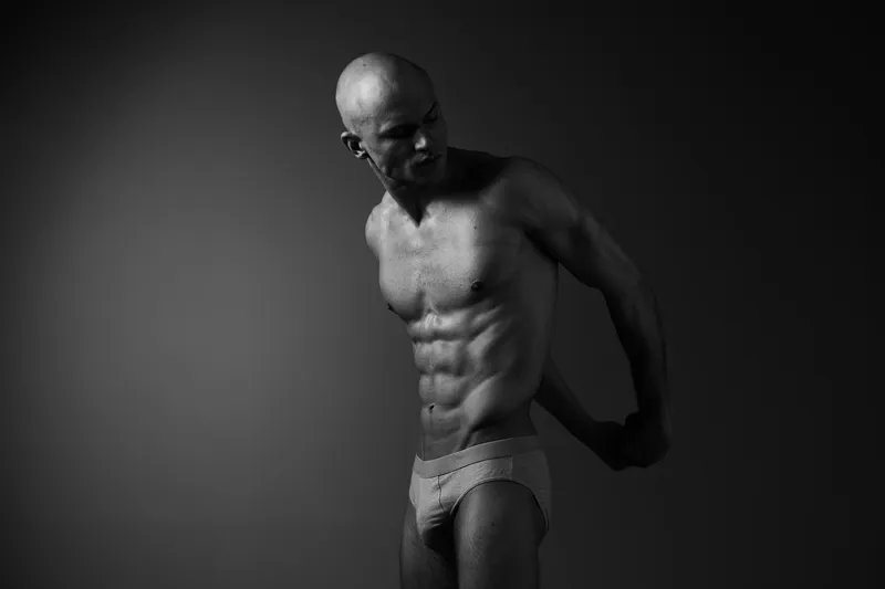 Black and white image of a muscular man posing confidently, showcasing strength and fitness with dramatic lighting highlighting his physique.