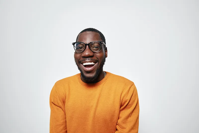 African American man with dark skin wearing glasses and orange sweater smiling happily on white background. Portrait of cheerful black male with beard and casual clothing.