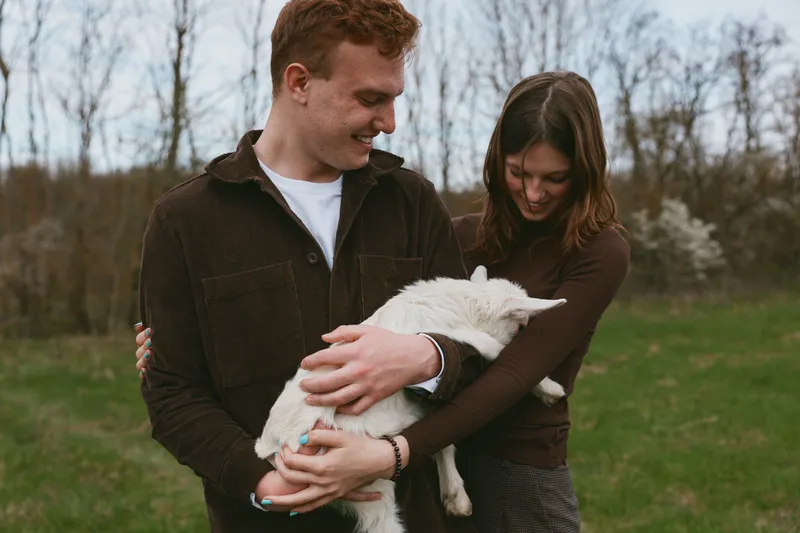 A couple outdoors with a gentle goat kid in their arms, on a sunlit farm meadow, capturing bonding, happiness, and rural family moments with animals in a natural lifestyle.