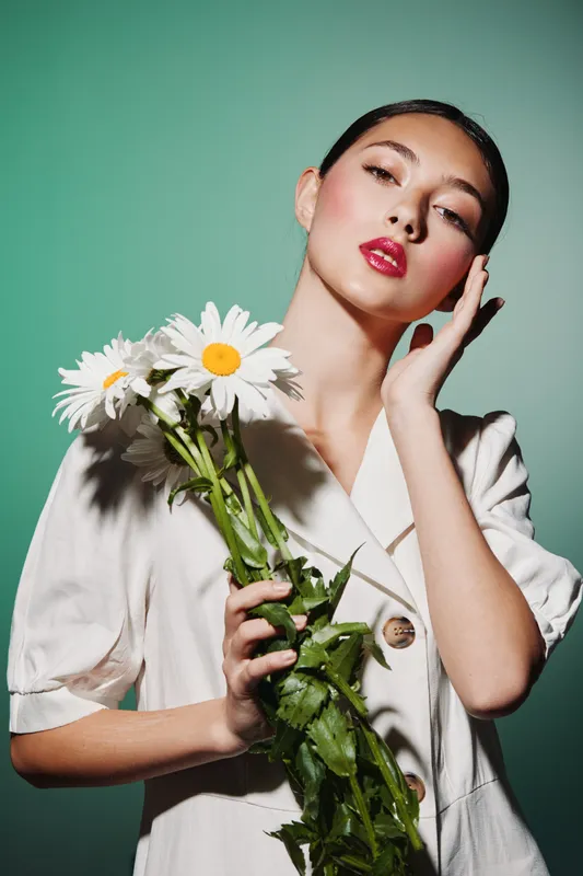 Young woman with glossy red lips holds white daisies against teal background in a creamy jacket. Concept of beauty, fashion, fresh floral, and creative styled portrait photography.