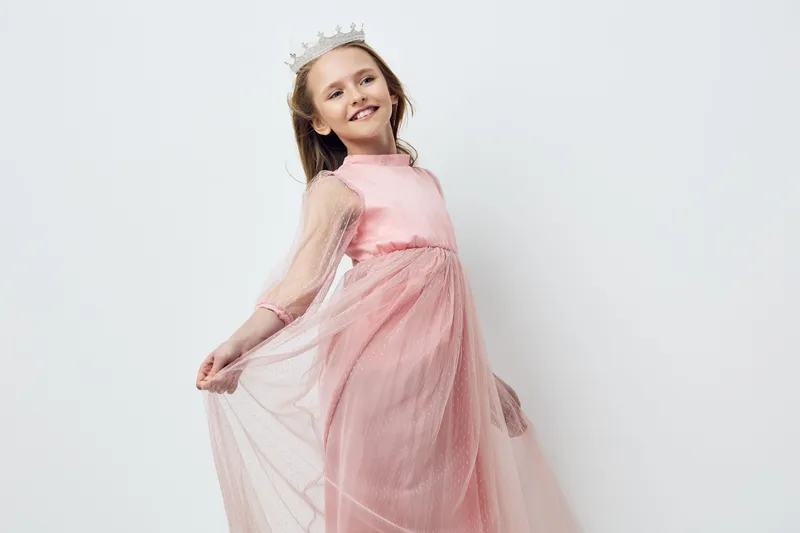 A joyful girl in a pink princess dress smiles while twirling, wearing a crown, against a light background, capturing the essence of childhood innocence and fantasy