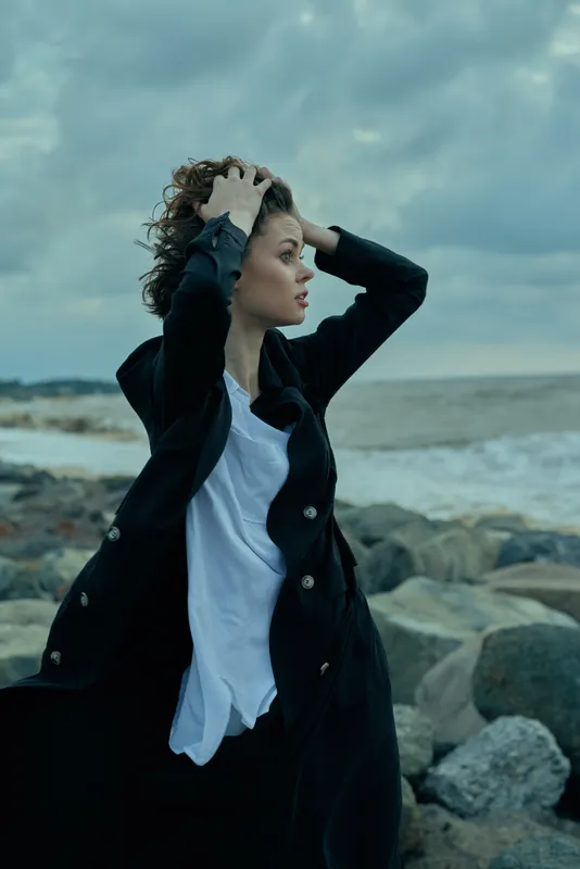 Coastal wind, woman in black coat and white shirt on windy shoreline with rocks, dramatic sky, windy scene, conveying mood, resilience, outdoor fashion and sea scenery