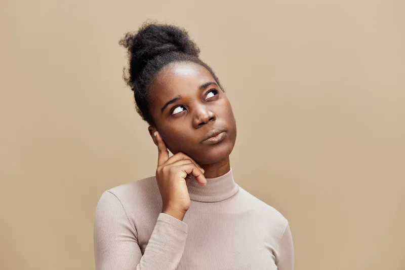 Thoughtful young Black woman with dark curly hair in a beige turtleneck contemplating on a light brown background, conveying curiosity and introspection
