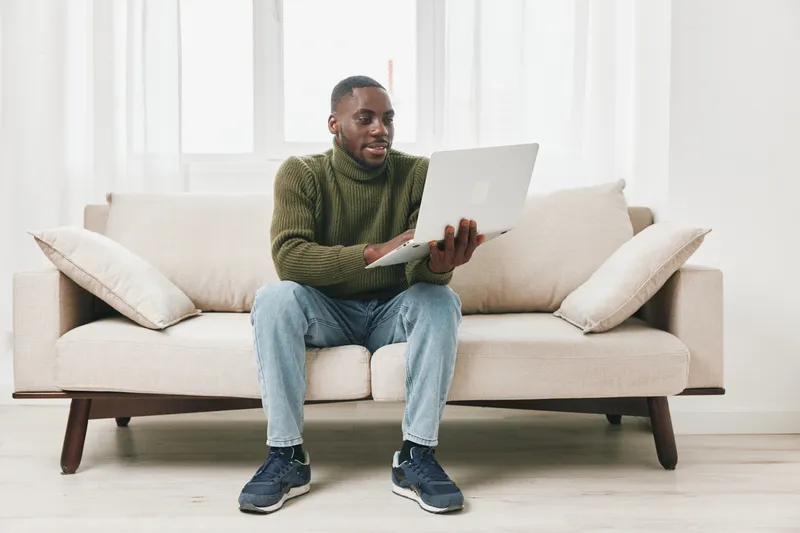 A young African American man engaging with a laptop on a cozy sofa, showcasing a modern and casual lifestyle in a bright indoor setting