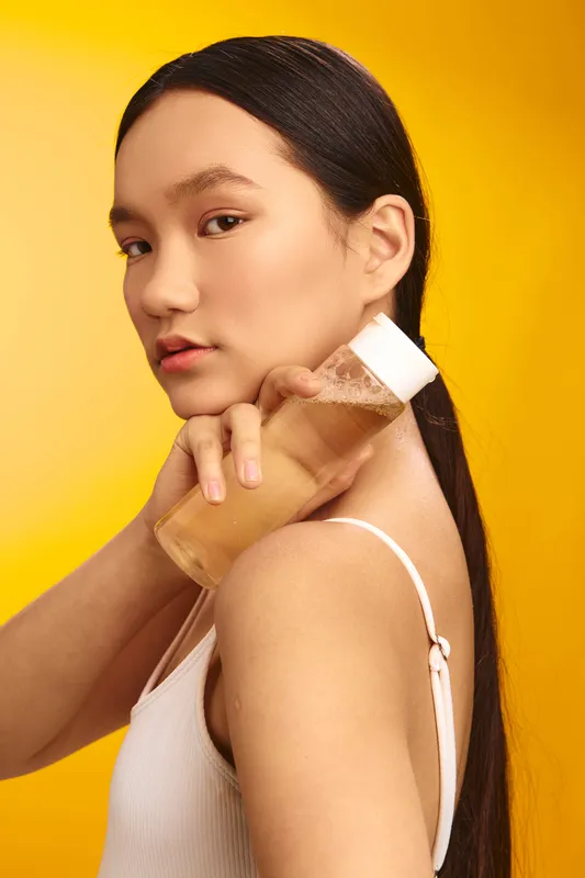 Asian woman stands against a bright yellow background, holding a cosmetic bottle. She exudes confidence and poise, creating a fresh, modern beauty moment that's crisp, stylish, and inviting.