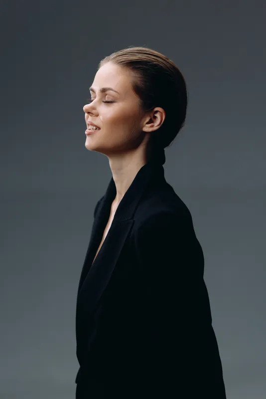 Profile of peaceful woman with closed eyes wearing black jacket in soft light against dark background