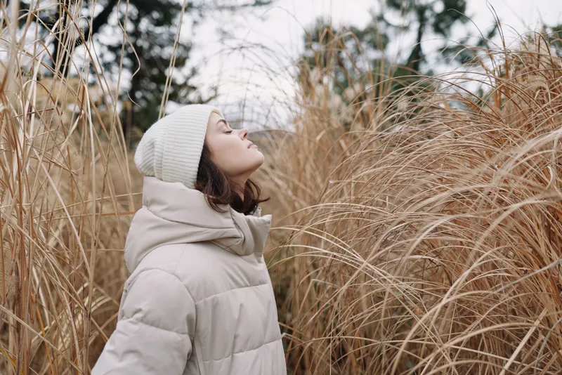 Woman in a warm coat and beanie stands among tall grasses, looking upward with contemplative calm in a winter outdoor nature scene.