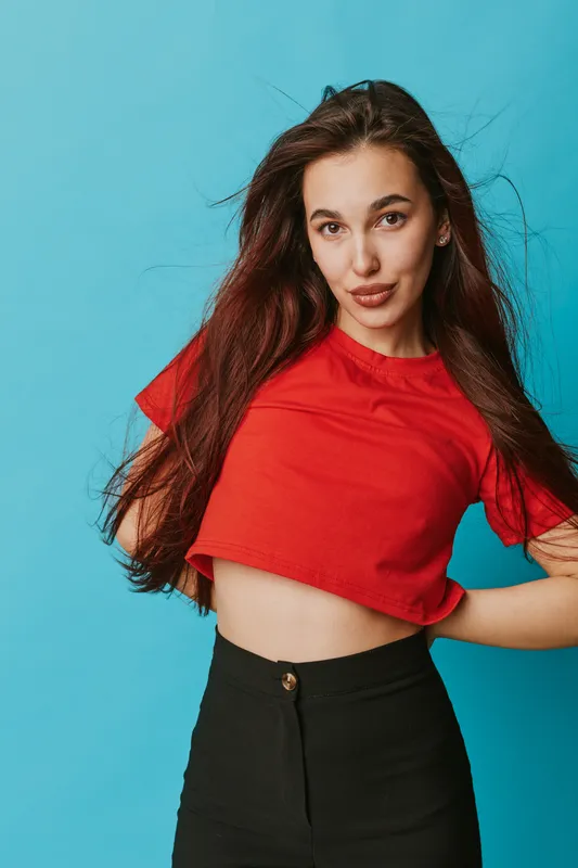 Young woman with long hair wearing red crop top and black pants posing confidently against blue background. Casual fashion, beauty and style portrait with natural makeup and relaxed expression.
