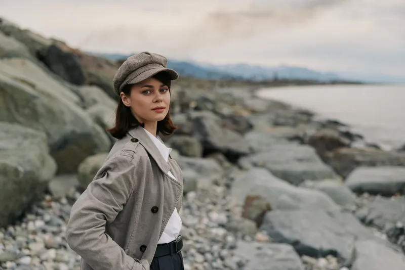 Young woman in vintage outfit stands on rocky beach looking at camera with calm expression. Overcast sky and distant mountains create moody coastal atmosphere.